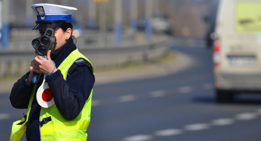 Policjantka ruchu drogowego (widoczna żółta kamizelka, biała czapka) mierzy prędkość ręcznym miernikiem. W tle - ruch uliczny / fot. KWP w Krakowie